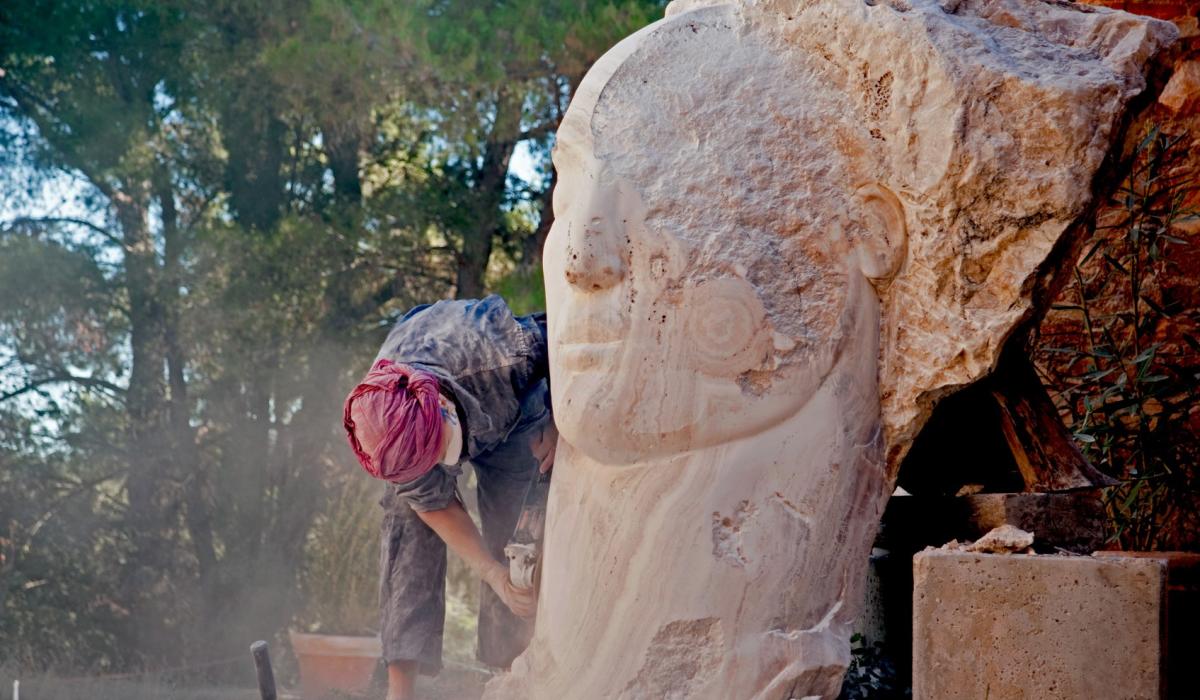 A sculptor works on a carved face emerging from a stone larger than her