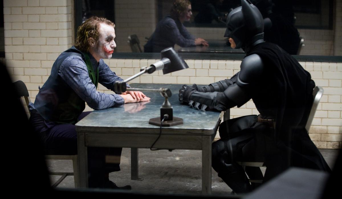 The Joker and Batman sit opposite each other in an interrogation room.