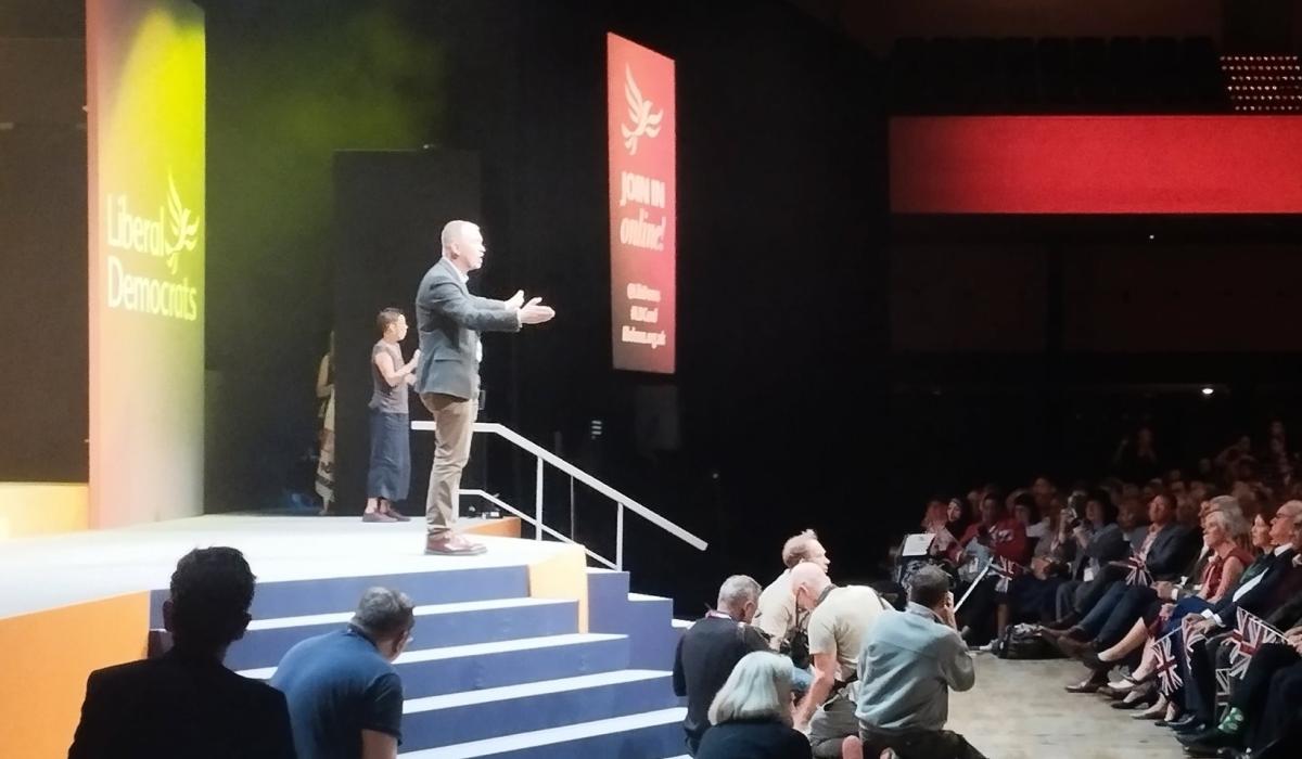 Tim Farron stands on a conference stage with arms wide