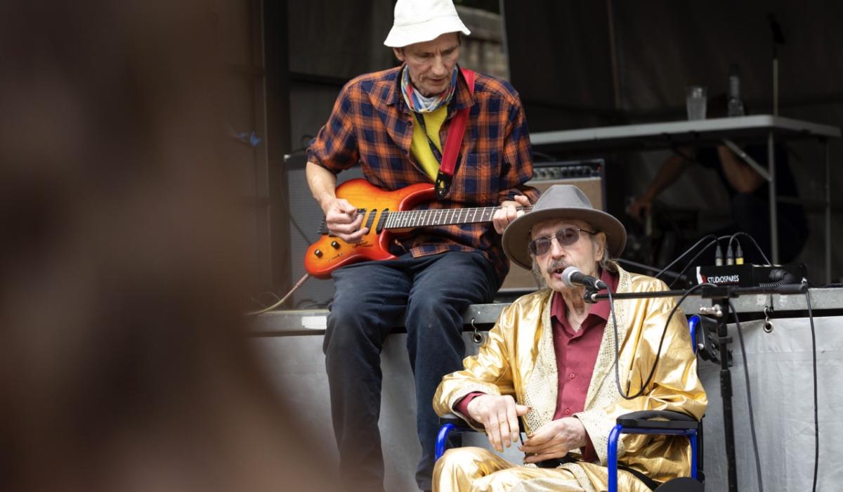 An elderly Elvis impersonator sits on a stage wearing a gold suit.