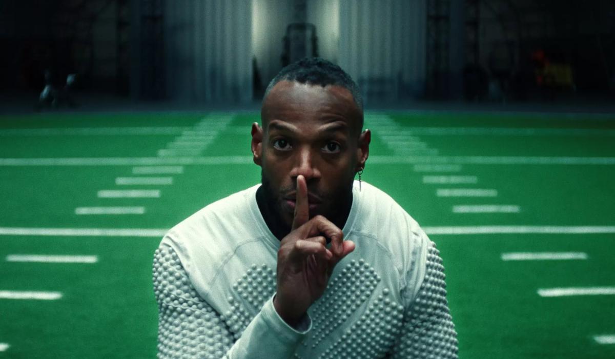 A player holding a finger to his lips stands in front of an indoor American football pitch.