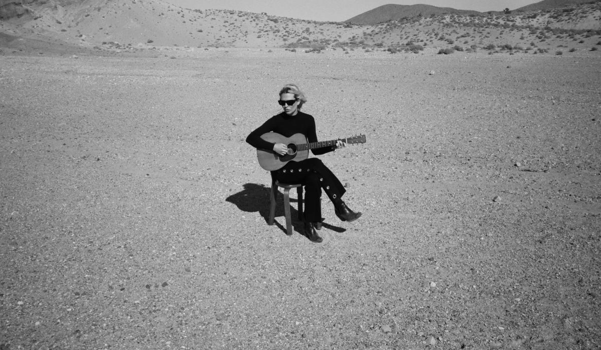A musician wearing black sits on a chair in a desert holding her guitar.