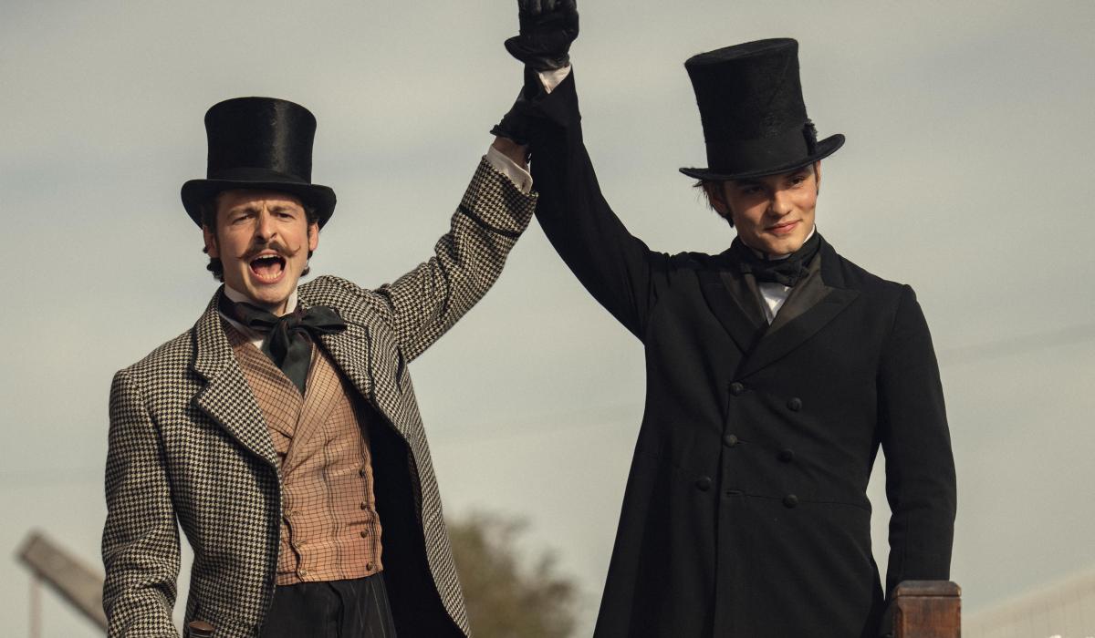 Dressed in Victorian clothes, two brothers raise their arms together.