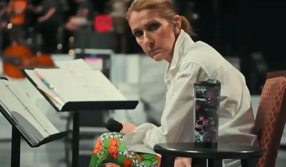 A seated Celine Dion, leans forward, head to the side, holding a mic.