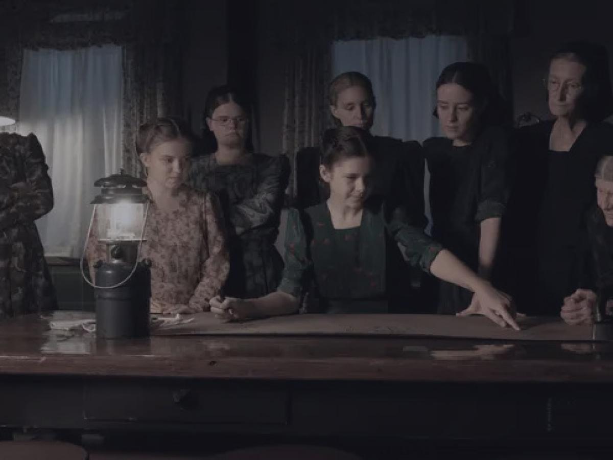 a group of women stand and sit around a table lit by a gas lamp.