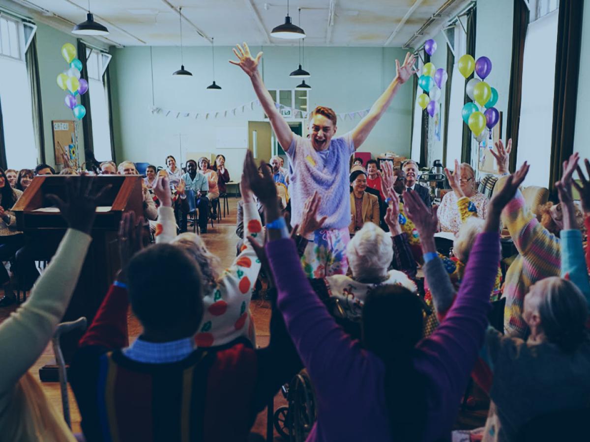 In a hall decorated for a celebration a person stands in front of a seated group, all have their arms raised in celebration.