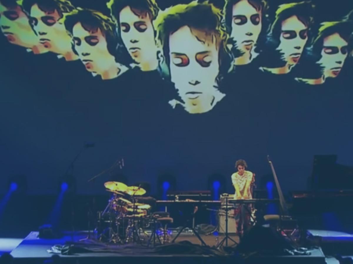 A muscian plays a keyboard on a concert stage surrounded by instruments, while multiple images of his face are projected behind him.