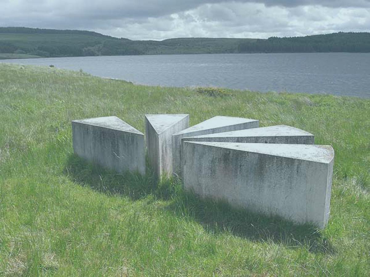 A set of low concrere blocks in the shape of a map's viewpoint symbol sit beside a lake.