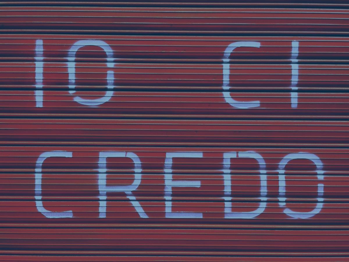 A red shutter door bears a large painted message reading 'io ci credo'