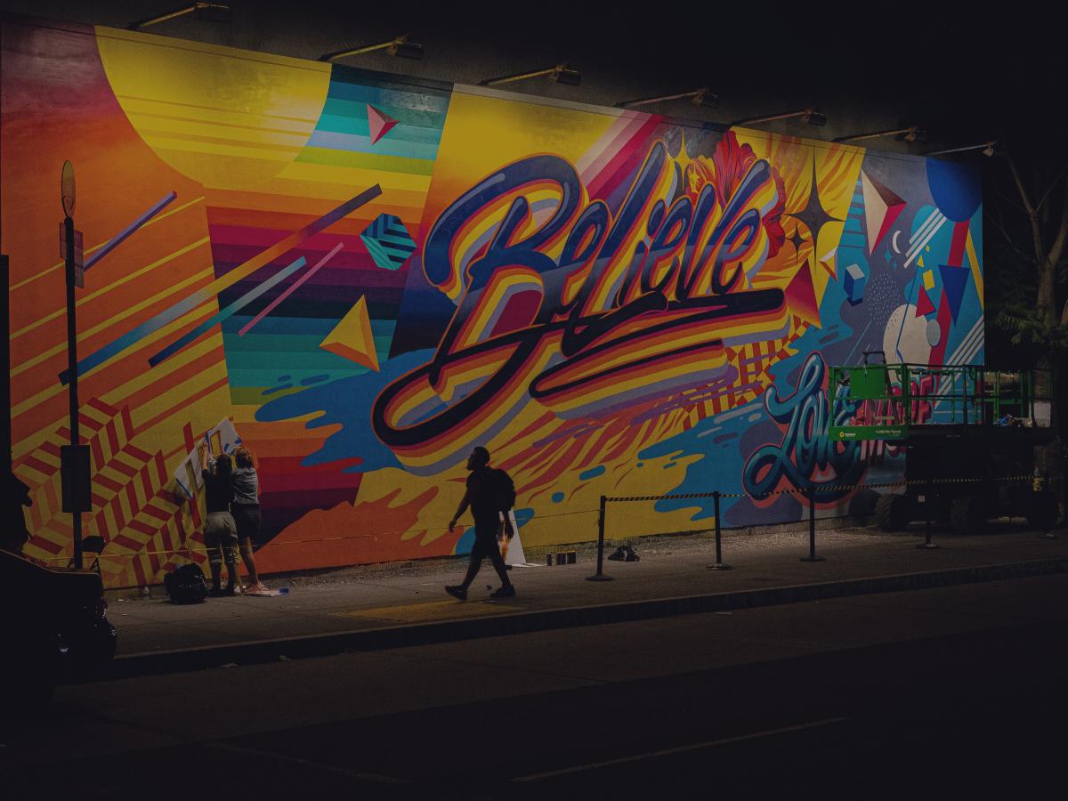 A person walks past a multi-coloured wall of graffiti with the word 'believe' in the centre of it.