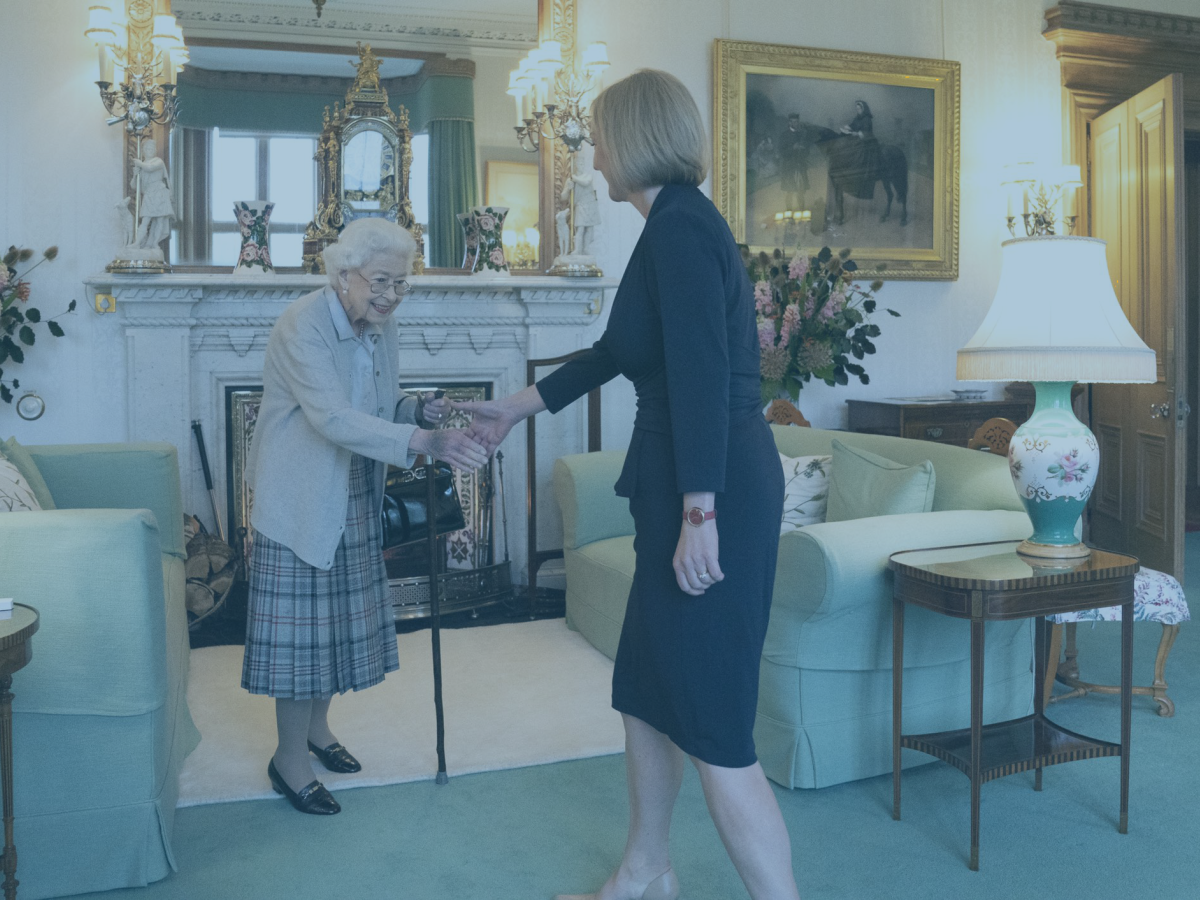 A frail old lady, the late Queen, rises from a sofa to shake hands with an approaching woman.