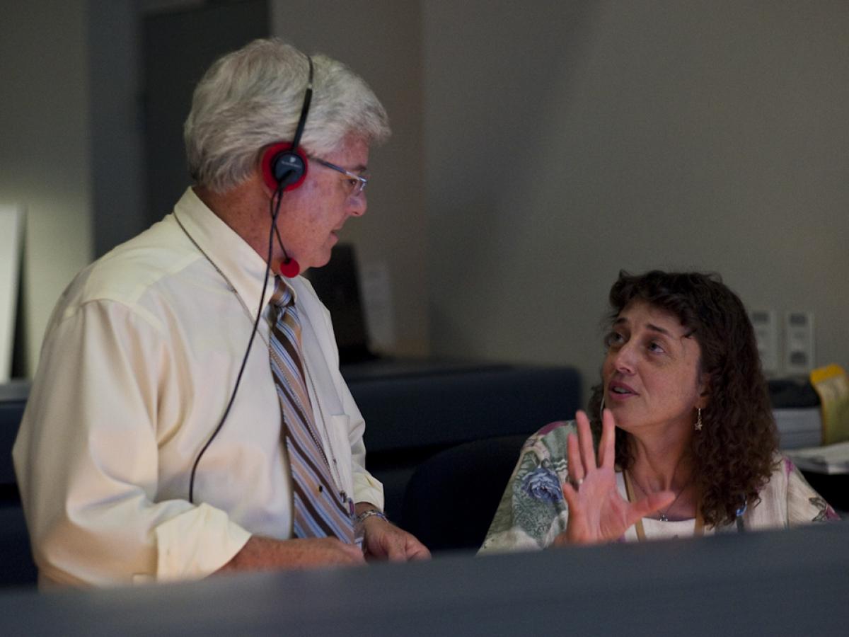 Two scientists talk to each other at console in a space mission control room.