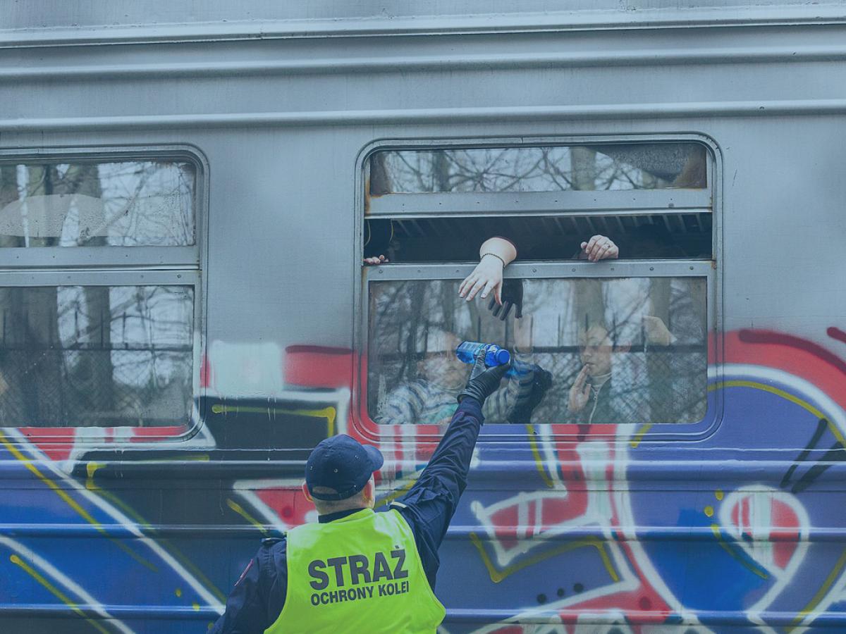 A helper in a yellow vest reaches up to a open train carraige window while offering a bottle. The side of the carraige is covered in graffiti.