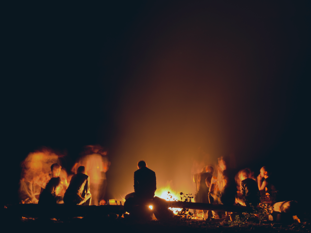a group of people crowd round a campfire backlighting them in silhouette.