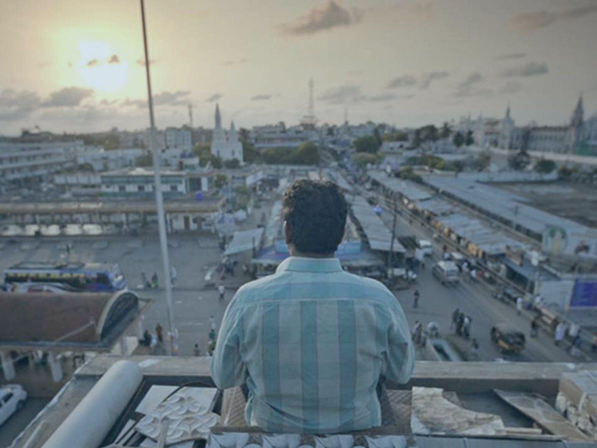 A man sits on the top of a building and looks out over the view of an Indian city as the sun sets.