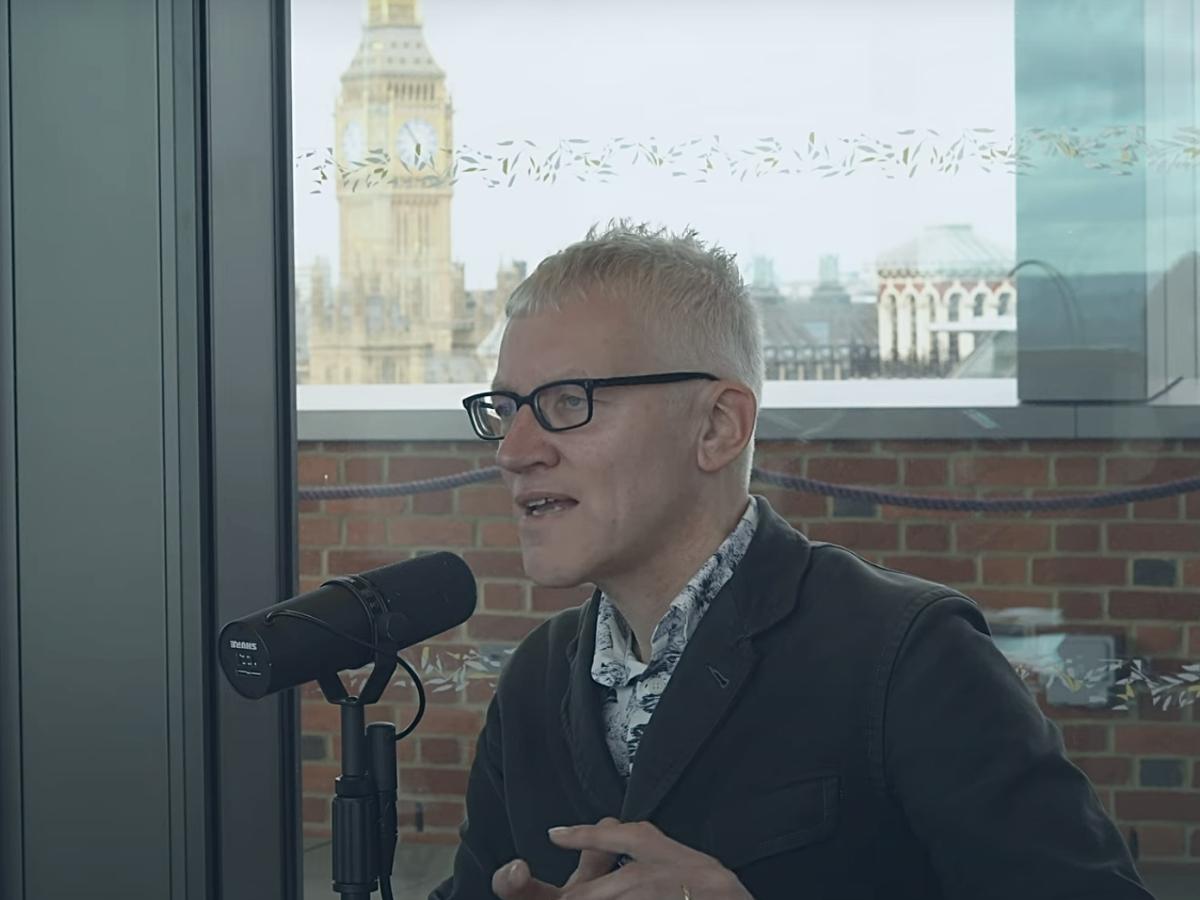 A man sits at a table speaking into a microphone but looks into the distance. Behind him, through the window and beyond a wall is Big Ben