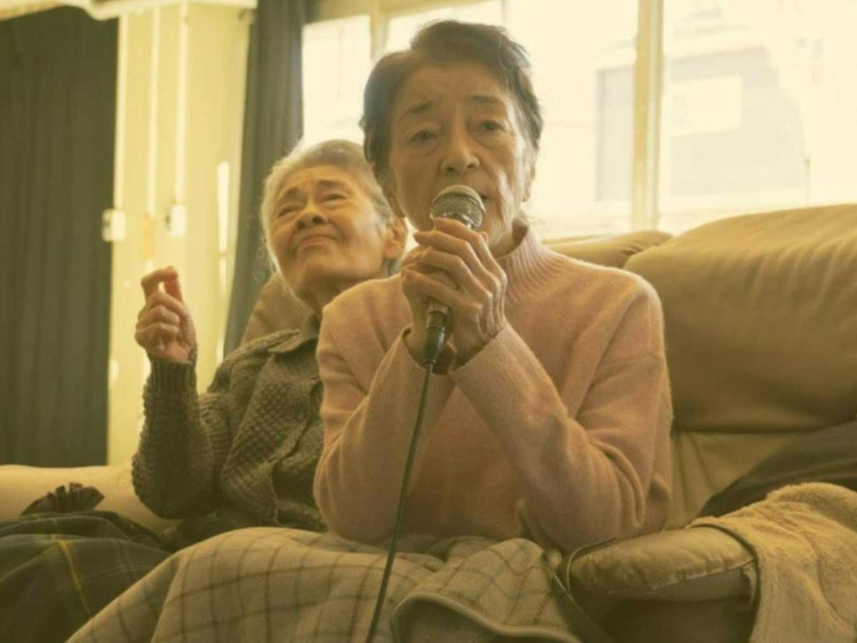In a retirement home, a older person sings karaoke while the person behind waves a hand.