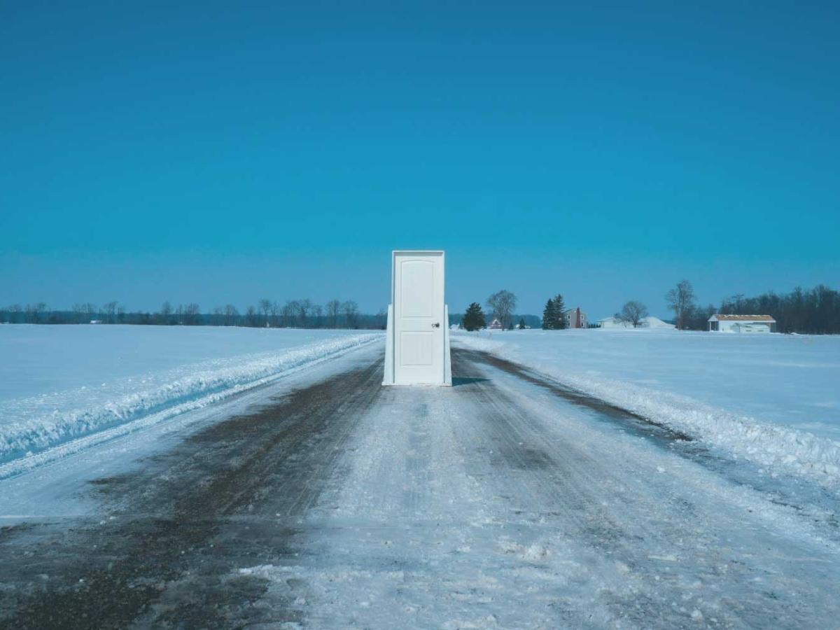A theatrical set door stands in the middle of a snowploughed road between fields of snow under a blue sky.