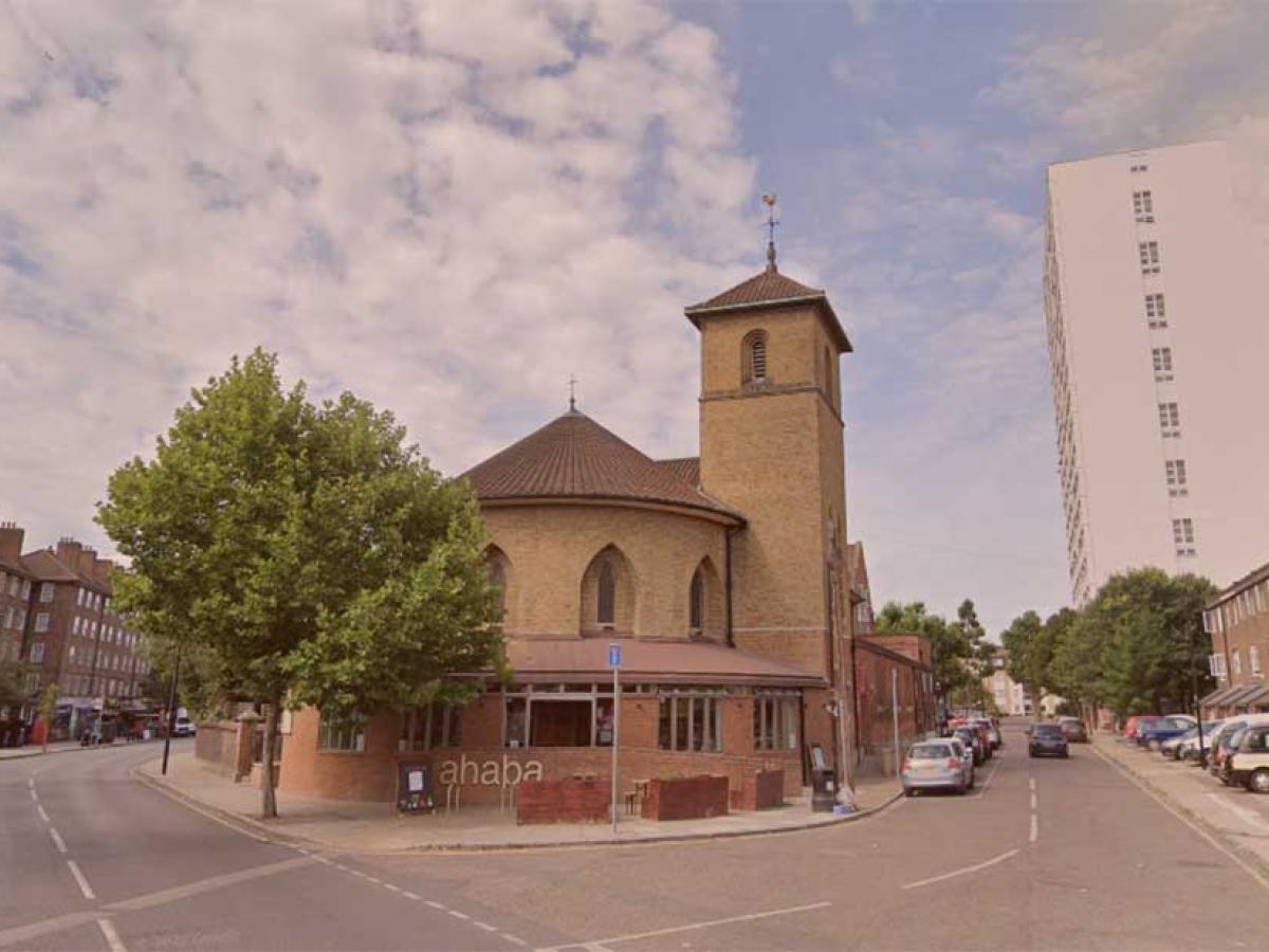 A neo-Romanesque church sits at the acute corner of two roads. To its side a tower block rises over a row of low-rise flats.