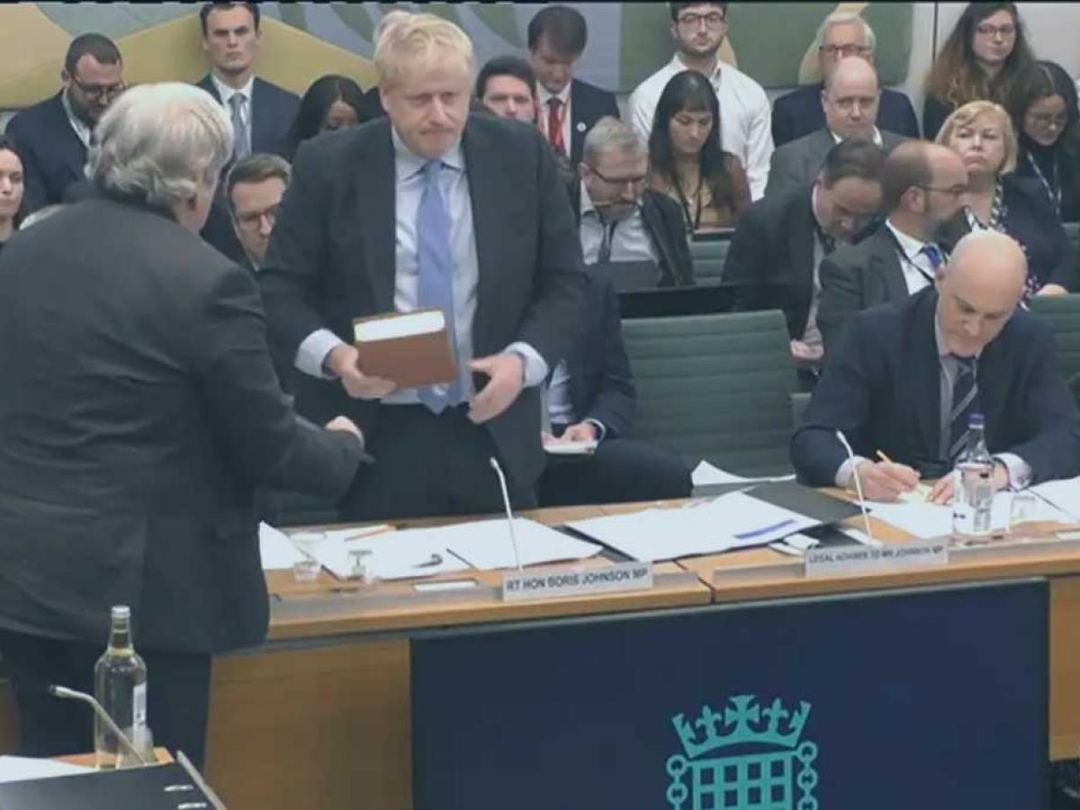 A politican stands holding a bible, in front of a committee room table. Behind him an audience waits expectantly