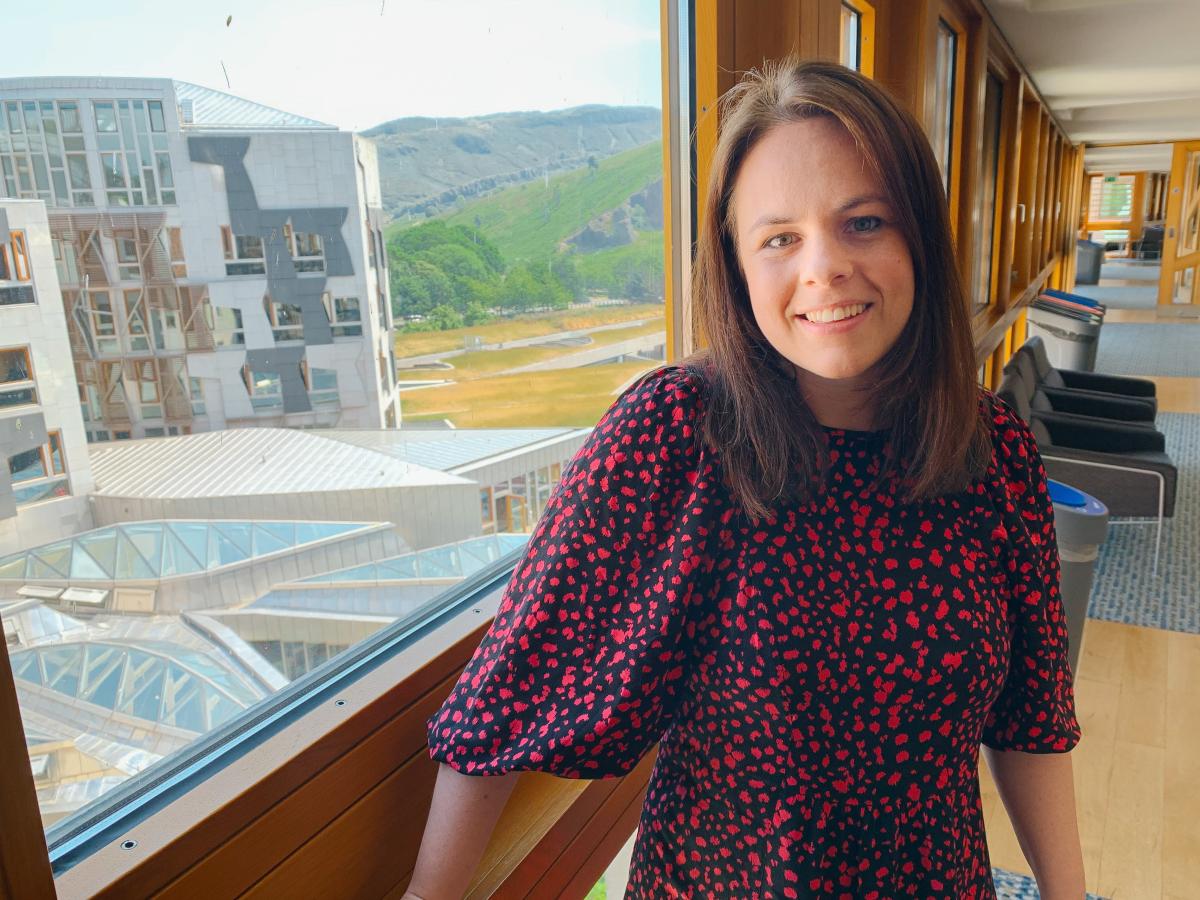 A women stands beside in a corridor beside a large window through which a wing of a building and a distant hillside can be see,