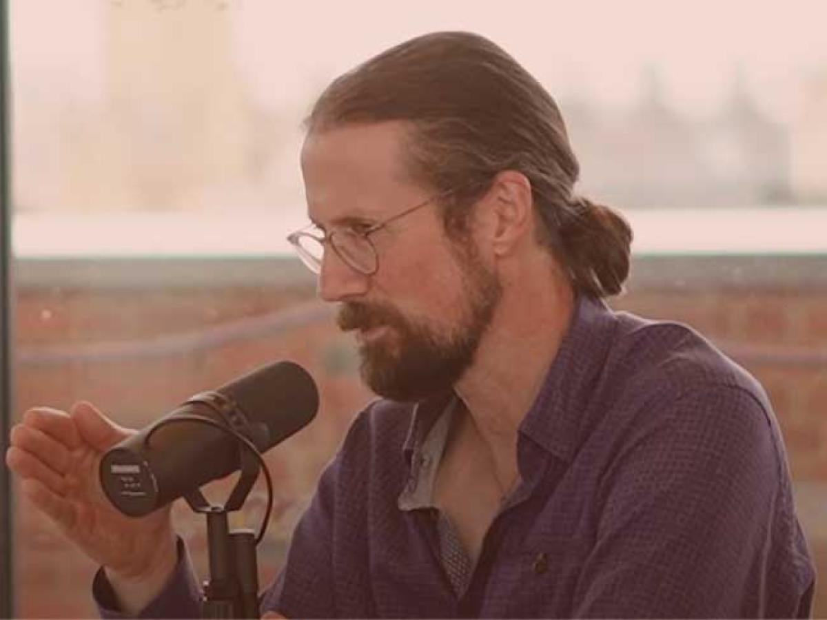 A sitting man speaks into a microphone will gesturing with one hand