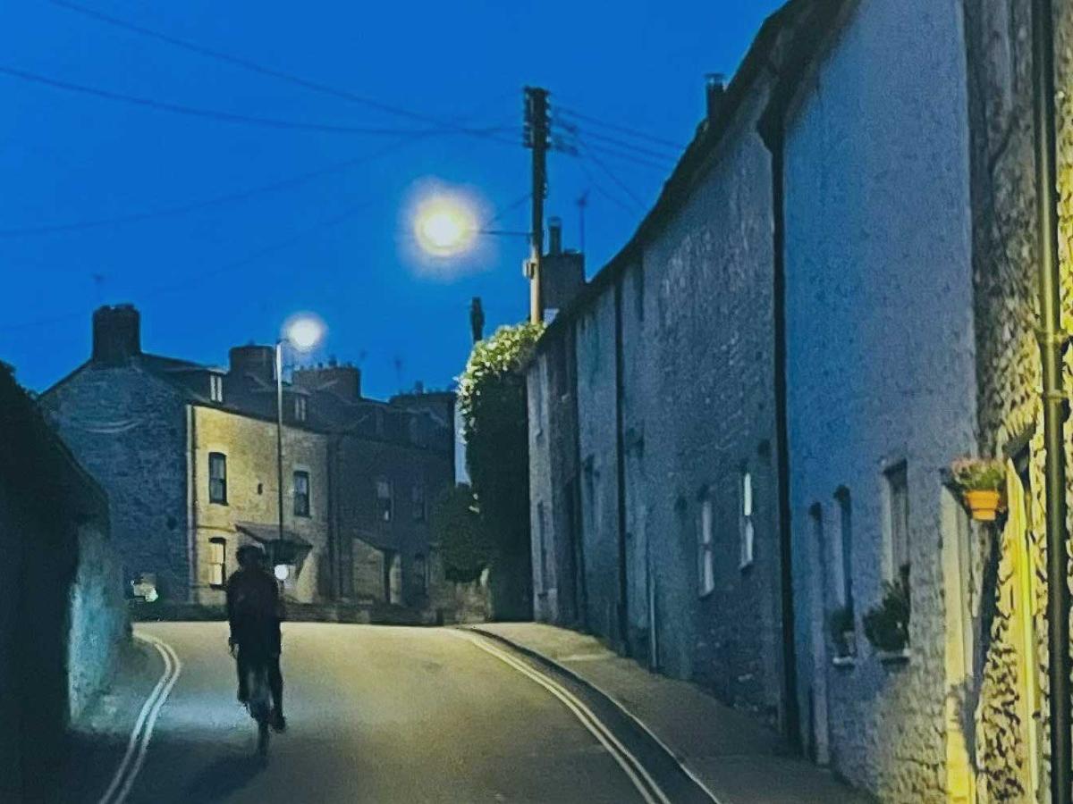 A cyclist ascends a village street between stone-built houses as twilight turns to night