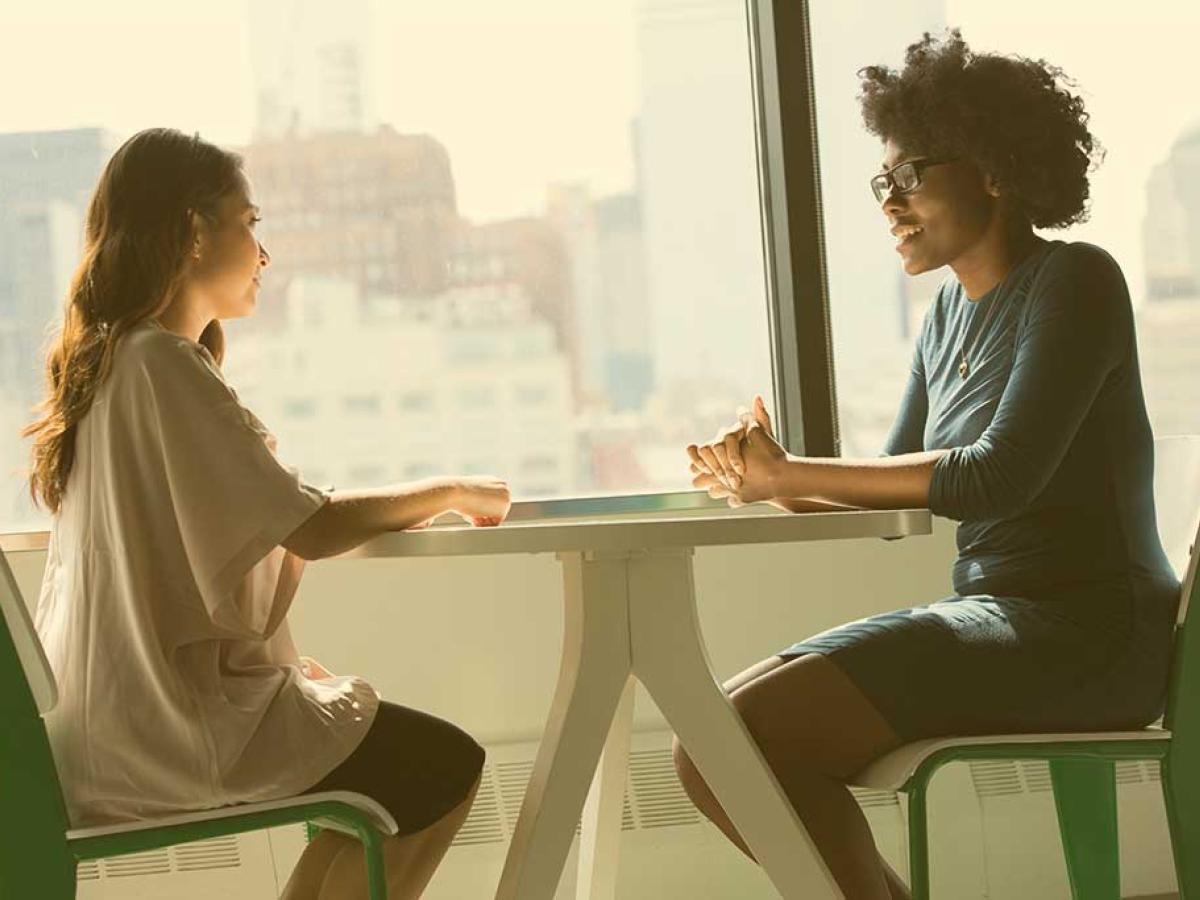 Two people sit at a table with their hands resting on top of it. One speaks to the other