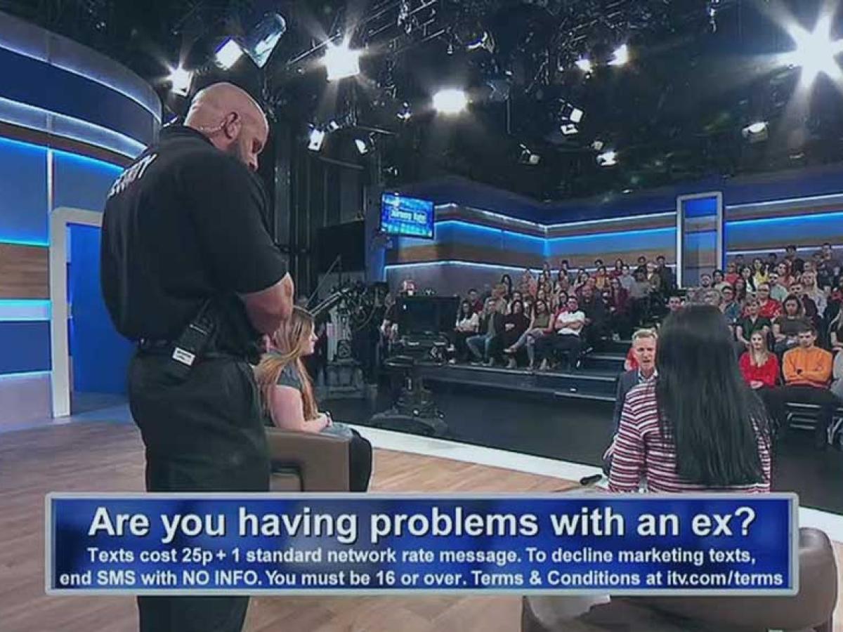 A view across a talk show TV set showing a security guard standing between two guest while the host talks to them. An audience looks on.