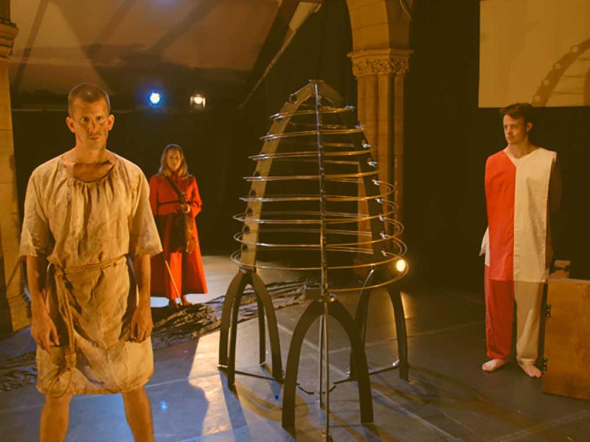 Three actors stand on a stage, in costume, surrounding a metal conical structure.