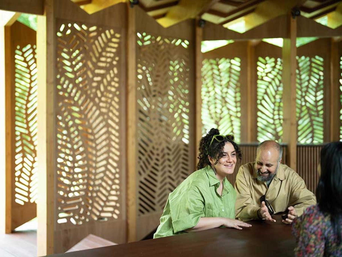 Around a table, against a backdrop of fret-cut wood, three people talk and listen to each other with great interest.