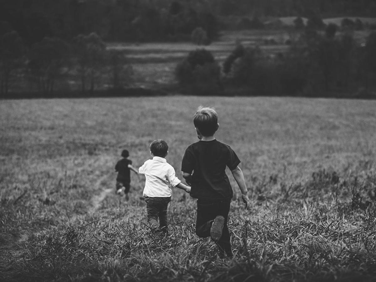 Three young children run away into the distance down across a field of long grass.