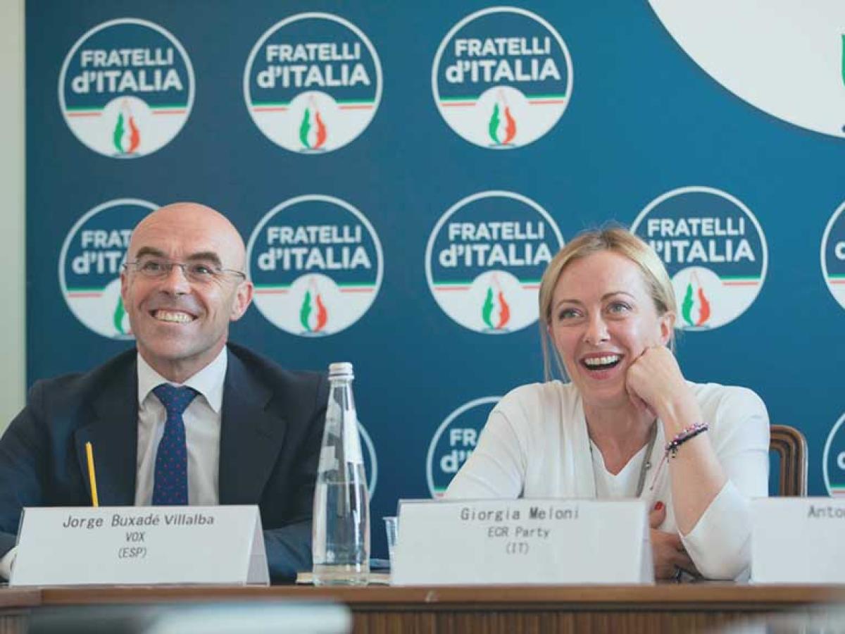 two politician site at a press conference desk and laugh, behind them is a backdrop of the political party's logo.