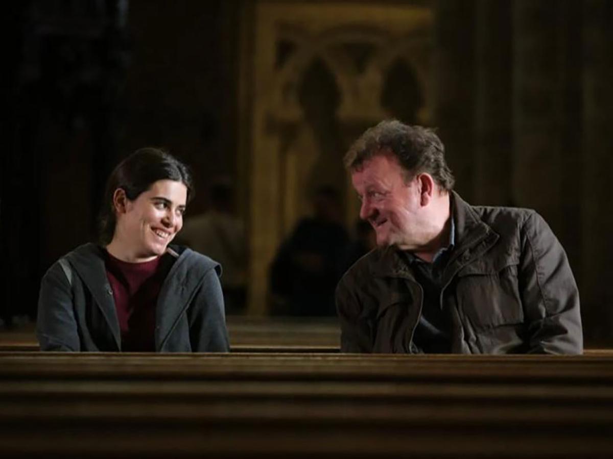 A man and a woman sit in a cathedral pew and incline their heads towards each other.