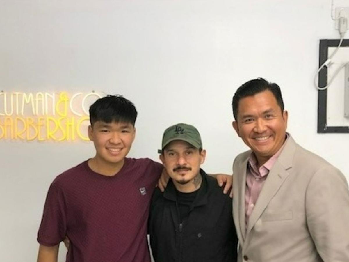 A barber stands between two clients, a father and son, a neon sign shines behind.