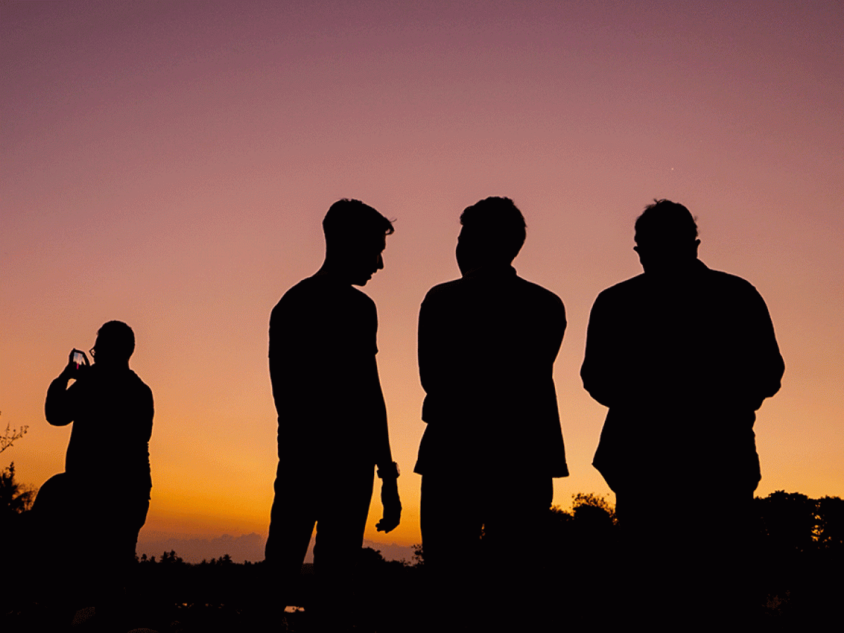 Four men stand silhoutted against a sunset, One stands apart on their phone.