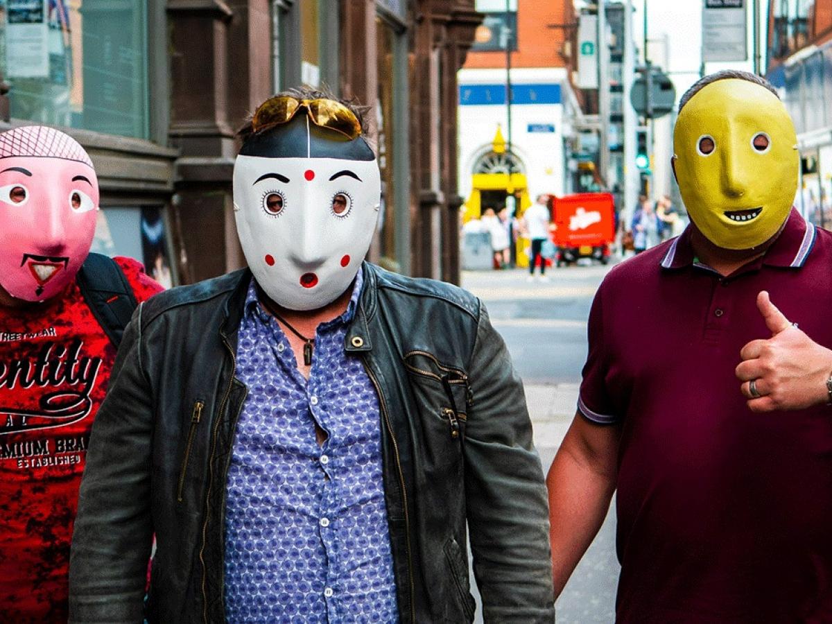 Three men wearing pink, spotty and yellow face masks stand in the street.