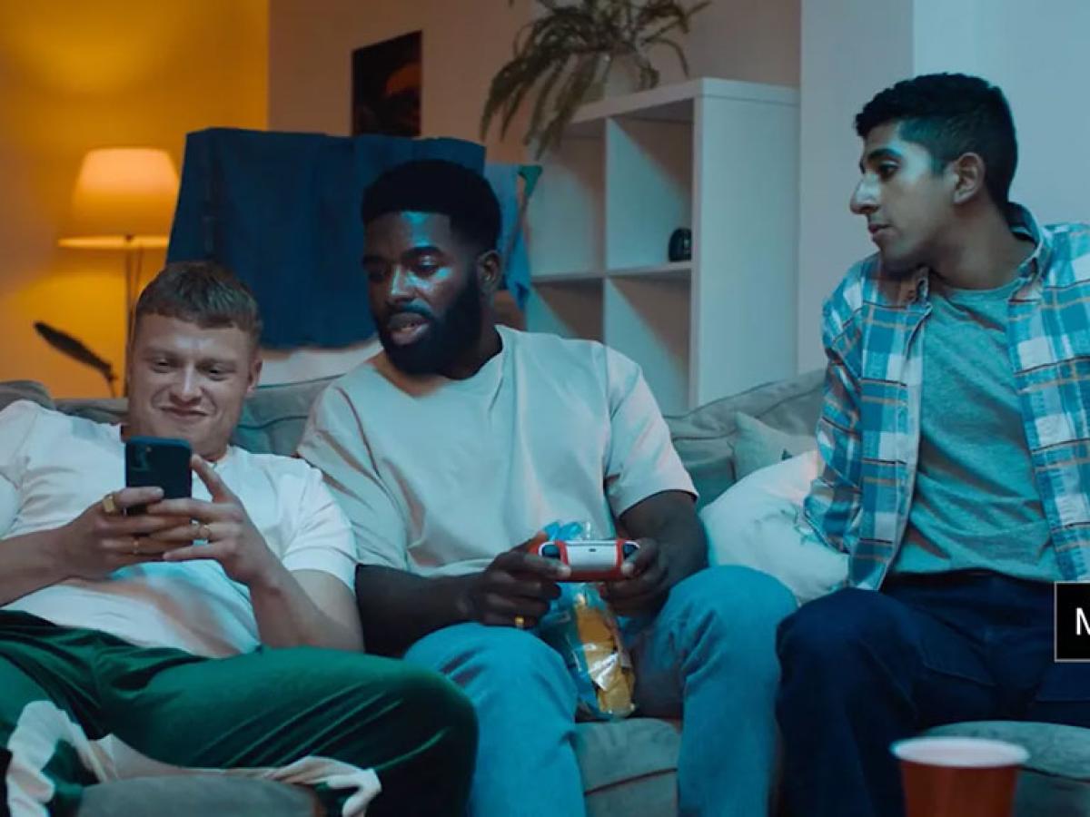 Three young men sit on a couch. One is leering at a phone while the others look on hesitantly
