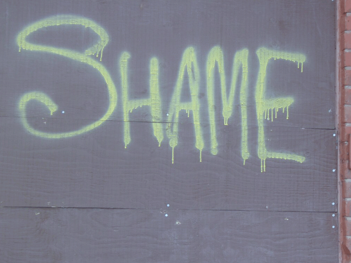 The word 'SHAME' spray painted onto a grey hoarding in lime green paint.