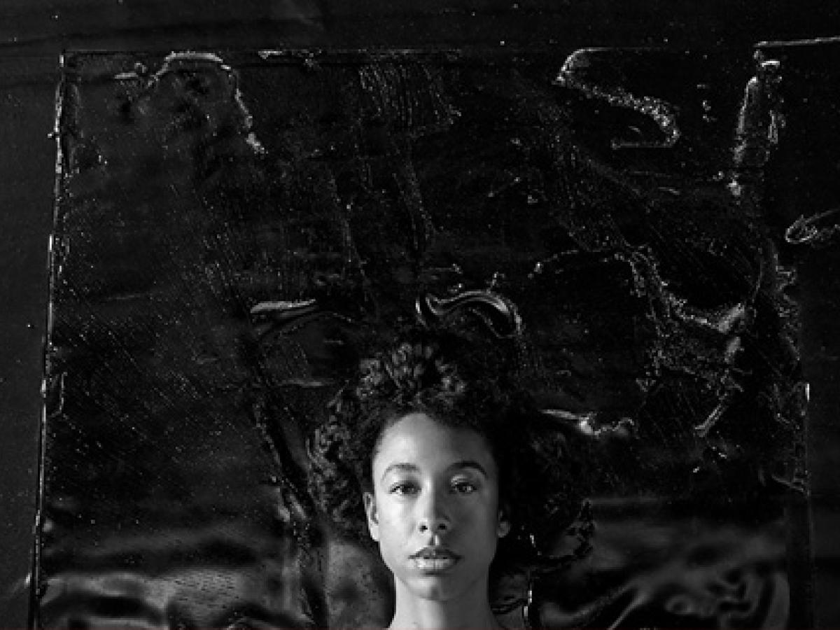 A black and white portrait of a young woman's head against a dark textured background.