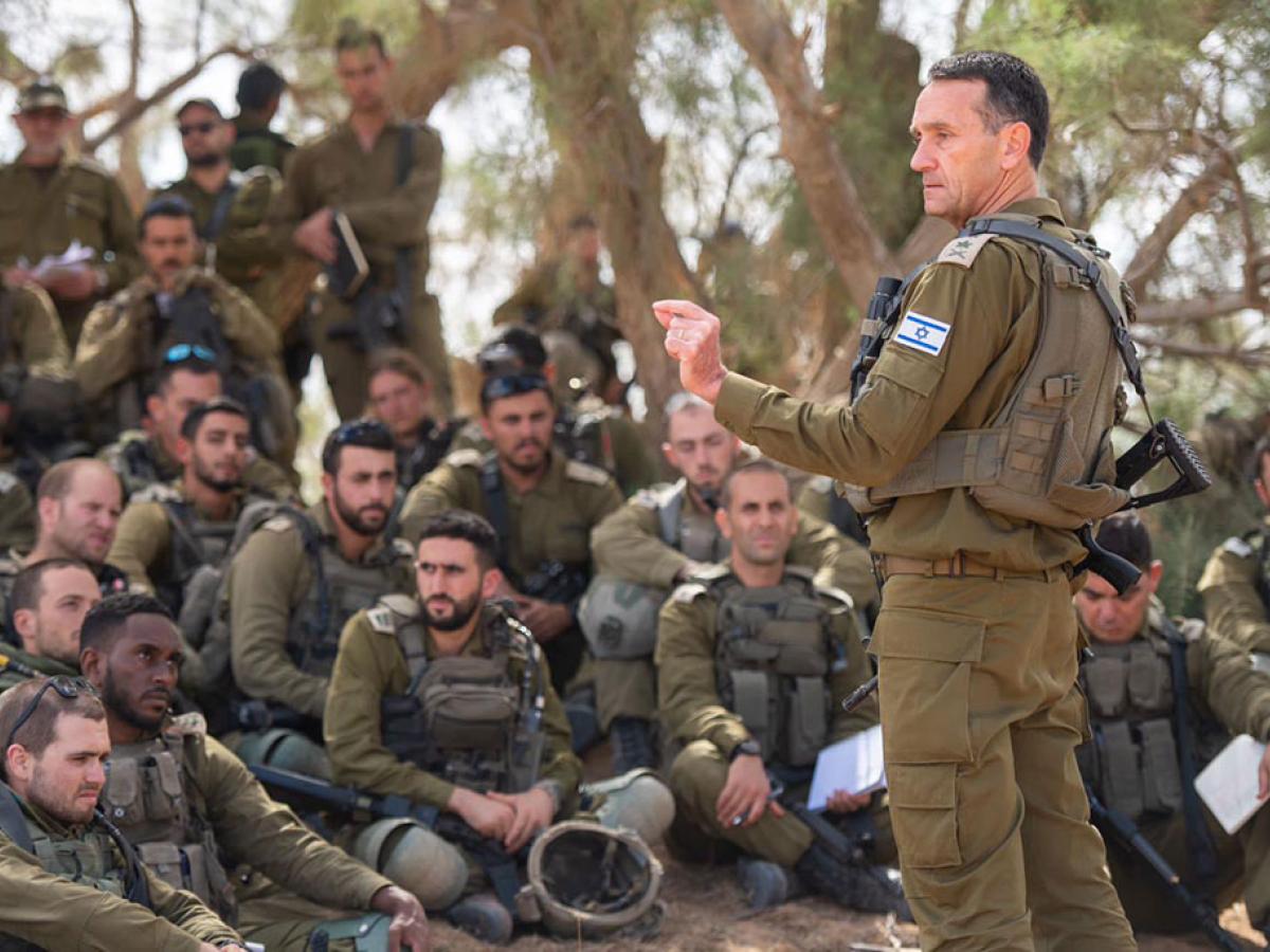 An army general in green fatigues stands and address a group of soldiers sitting, squatting and standing around him