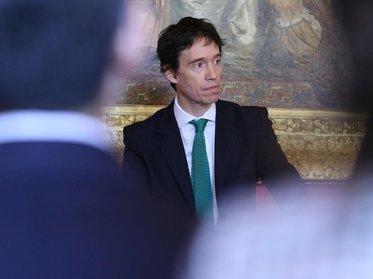 A suited politician stands looking pensive, framed by two out of focus audience members.