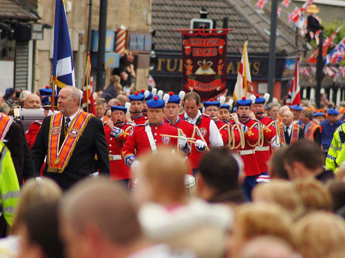 Across the heads of a roadside crowd, men wearing orange sashes and military band uniforms march along.