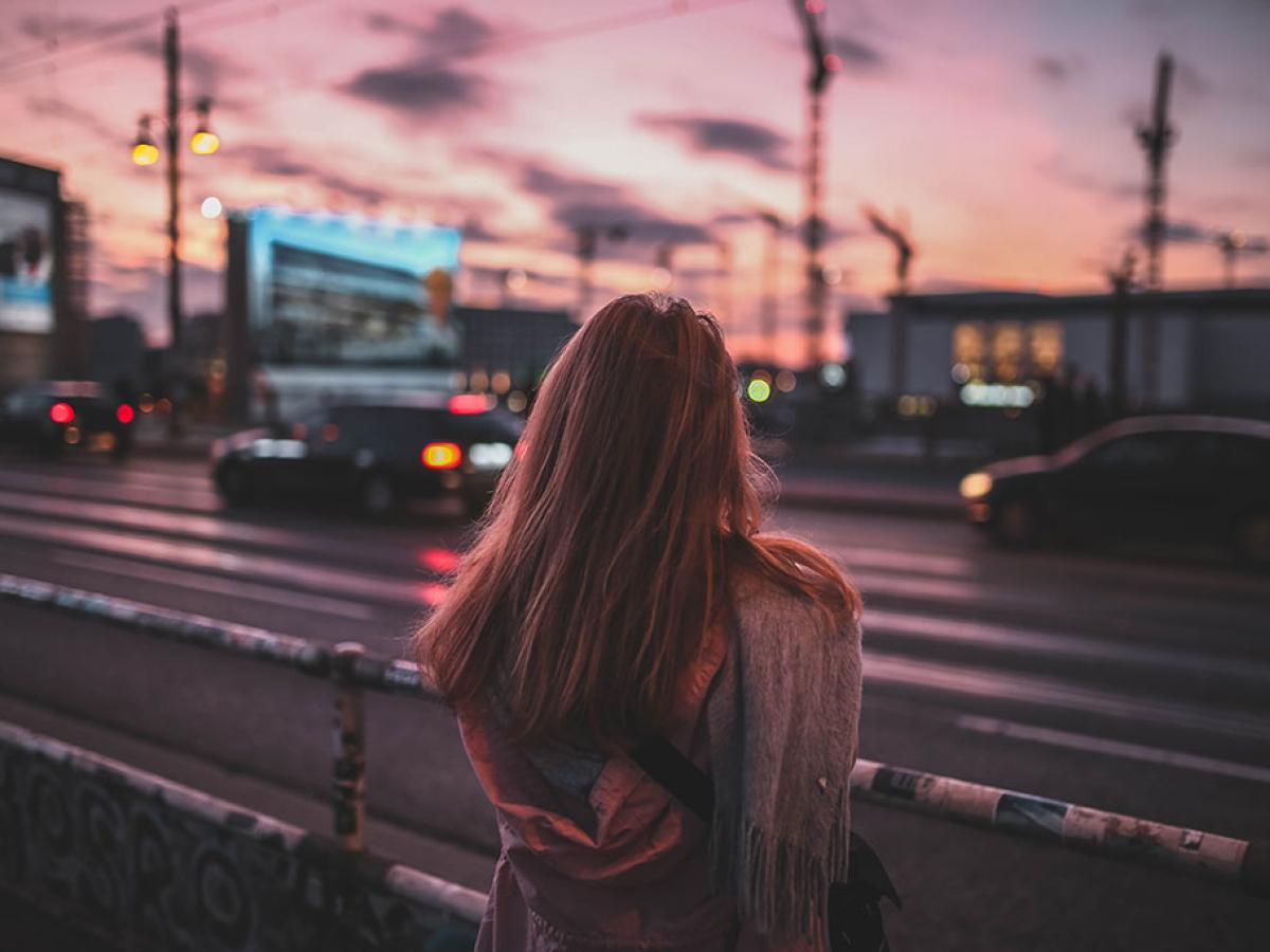 A woman stand beside a busy road with her back to the camera. There is a red sky behind her.