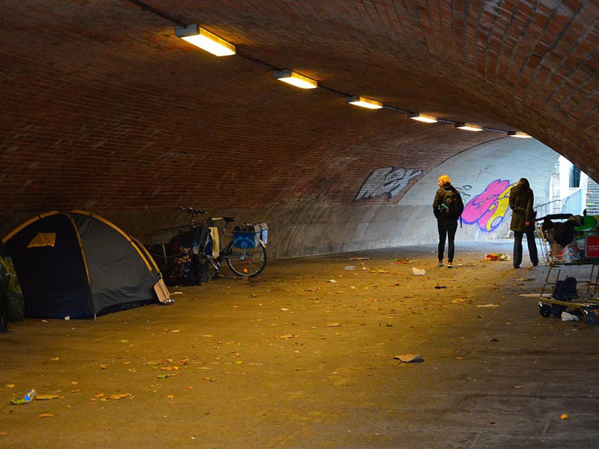 In an underpass a pedestrian passes and look at the tent of a homeless person.