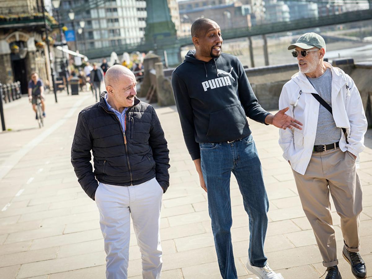 Three men walk down a path, the middle one talking and gesturing while the others listen.