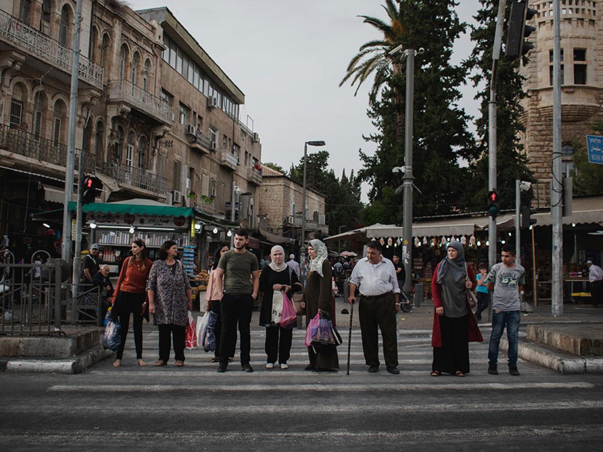 A line of people, some old, some young, wait to cross a road.