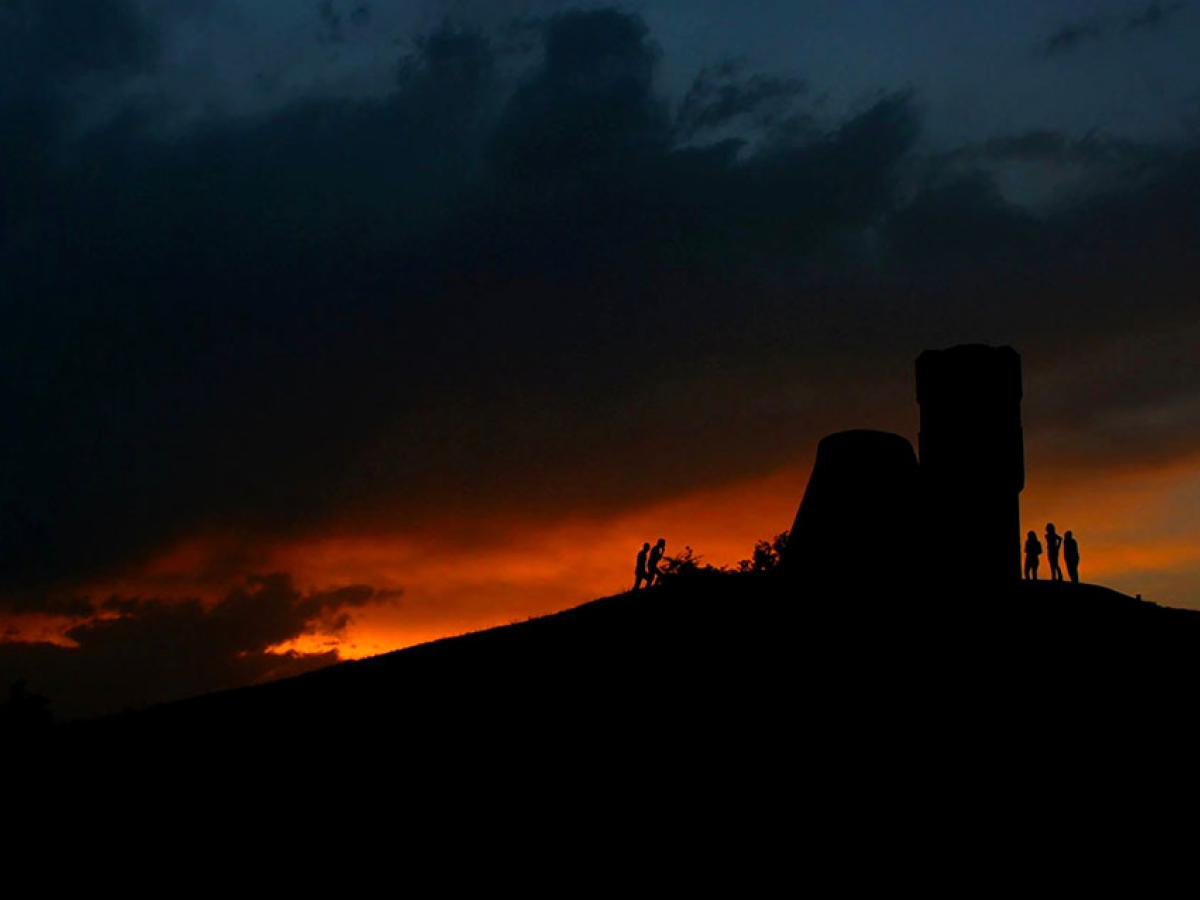 A sunset dramatically silhouette's a ruined tower and people at its base.