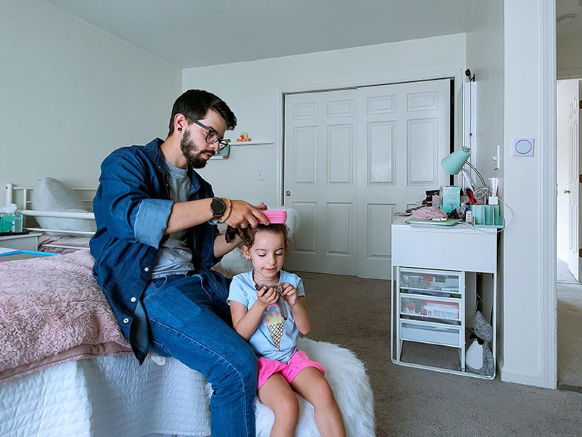 A father sits on a bed and fixes the hair of his daughter standing in front of him