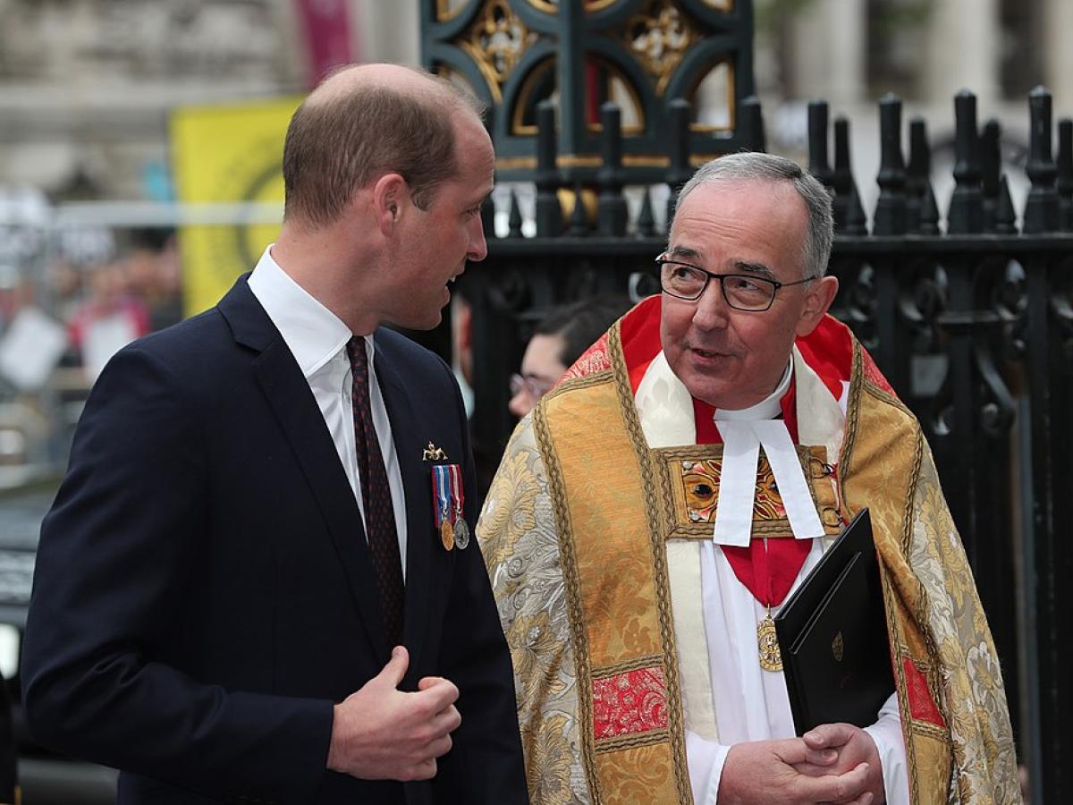 A young man wearing a dark suit talks to a minister wearing regalia.